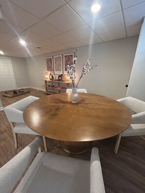 A dining area with a round wooden table, white upholstered chairs, a vase with branches, and a kitchen area in the background. - Mid-Century Modern Themed Ranch-Styled Surprise House (Delafield)