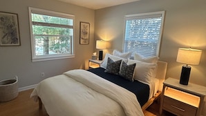 Quarto com uma cama grande, duas mesas de cabeceira com luminárias, uma luminária no teto e duas janelas com persianas.