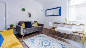 Un salon moderne avec un canapé gris, une table à manger avec des chaises, un plaid en fausse fourrure blanche et une grande fenêtre avec des stores.