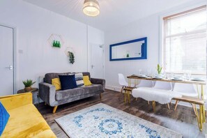 A modern living room with a grey sofa, a dining table with chairs, a white fur throw, and a large window with blinds. - Stylish | 4BR | 3Bath | Manchester Home (Eccles)