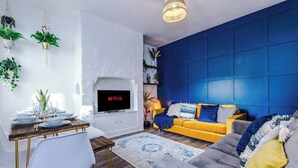 Un salon moderne avec un mur d’accent bleu, un canapé jaune, une cheminée et un coin repas avec une table dressée pour quatre personnes.