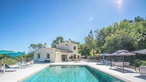 Zona de piscina con sillones de descanso y sombrillas, una casa al fondo y cielo azul despejado.