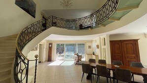 Un comedor amplio con una gran mesa de madera y sillas modernas, una escalera con barandillas de metal ornamentadas y vistas al exterior a través de puertas de vidrio.