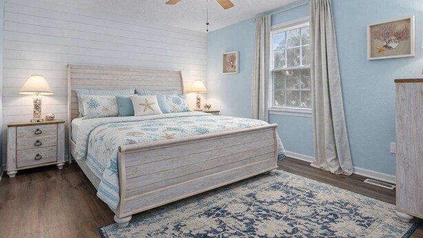 A bedroom with a wooden bed, two bedside tables with lamps, a ceiling fan, a window with curtains, and a framed picture on the wall. - 5BR Beach House in WW Crest - Sleep 12! (Wildwood)