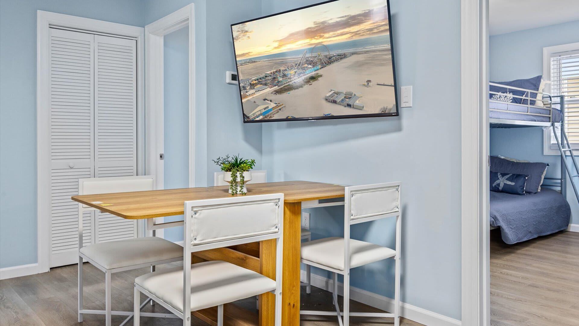 A room with a wooden dining table and chairs, a bunk bed with blue bedding, a TV mounted on the wall, and a potted plant on the table.