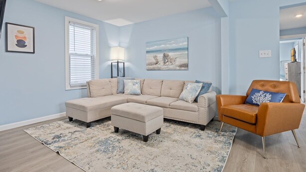 A living room with a sectional sofa, a chaise lounge, a rug, and a painting of a beach scene. - North WW 3BR 1st Floor - Steps to the Beach! (North Wildwood)