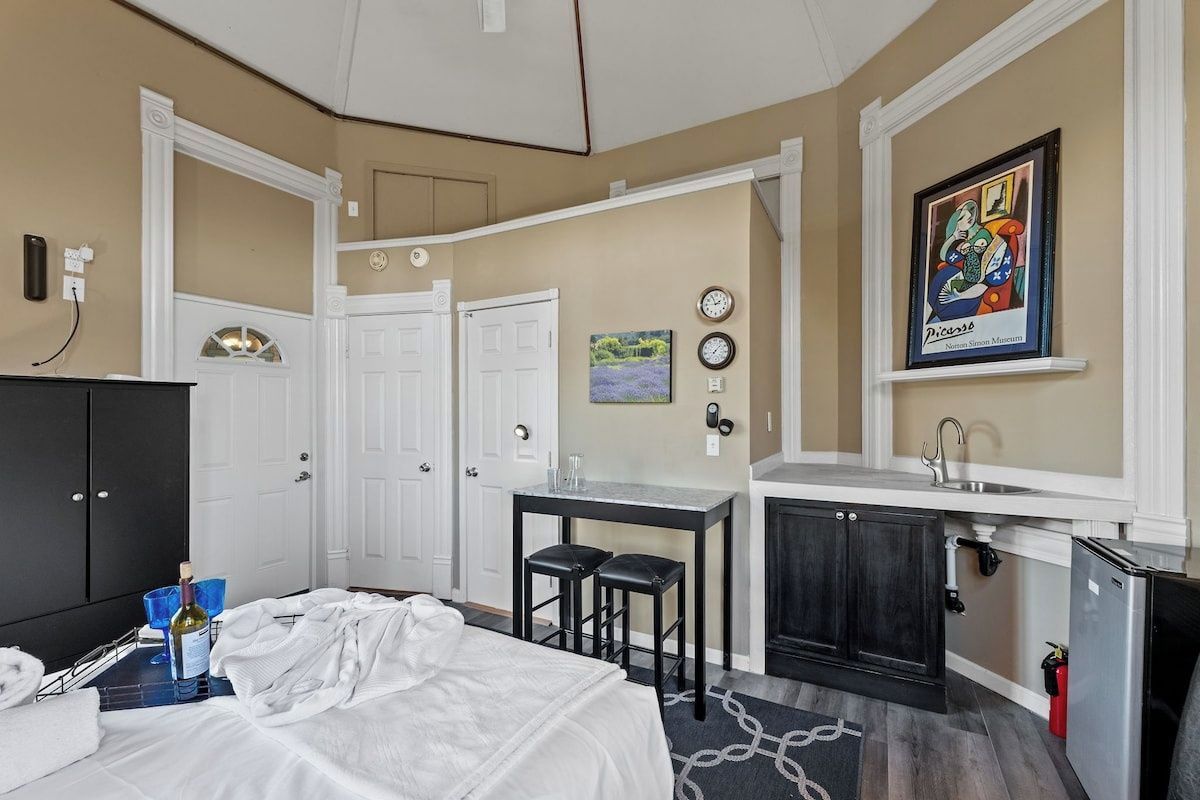 Une pièce de taille modeste, comprenant un lit, une petite kitchenette, une porte blanche et un tableau accroché au mur.