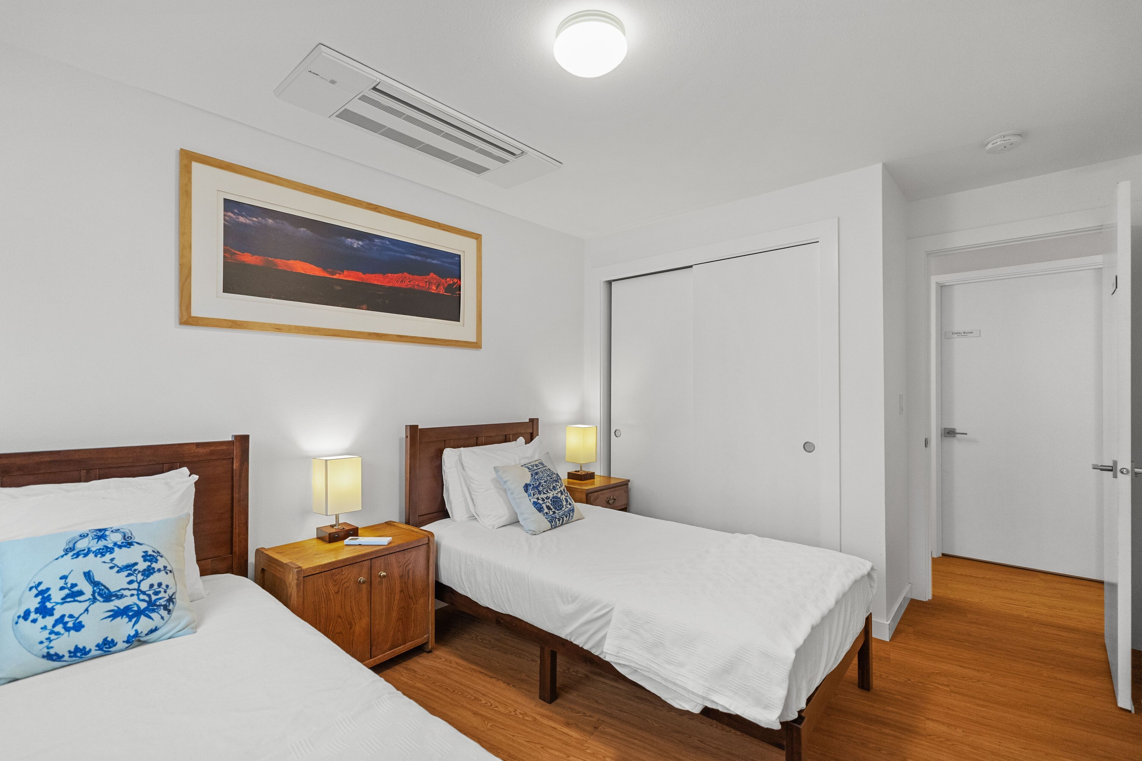 A hotel room with two single beds, wooden headboards, bedside tables with lamps, a framed picture on the wall, and a white door.
