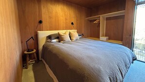 Un dormitorio con techo de madera, cama con cubrecama y almohadas grises, mesita de noche con lámpara y armario de madera.