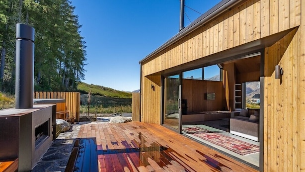 Una cabaña de madera moderna con terraza, chimenea y un amplio ventanal que da a una sala de estar.