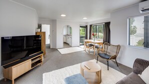 Ein modernes Wohnzimmer mit Flachbildfernseher, einer Holz-Unterhaltungseinheit, einem runden Couchtisch, einer Couch, einem Essbereich mit Tisch und Stühlen sowie einem großen Fenster mit Jalousien.