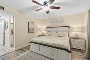 A bedroom with a bed, nightstands, a ceiling fan, and a welcome sign. - Sea Coast Gardens II 207 (New Smyrna Beach)