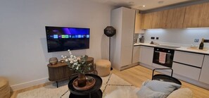 A modern living room with a flat-screen TV, a coffee table with a vase, a sofa, and a kitchen area with wooden cabinets and a countertop. - Open Mind Property – New Build 1-Bed w/ Parking (Bournemouth, BH5 1AE)