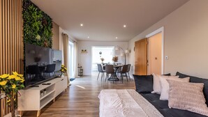 A modern living room with a flat-screen TV, a sofa, a coffee table, and a dining area in the background. - Open Mind Property- 6BR Home, Hot Tub & Free Parking (Fareham , PO15 5RS)