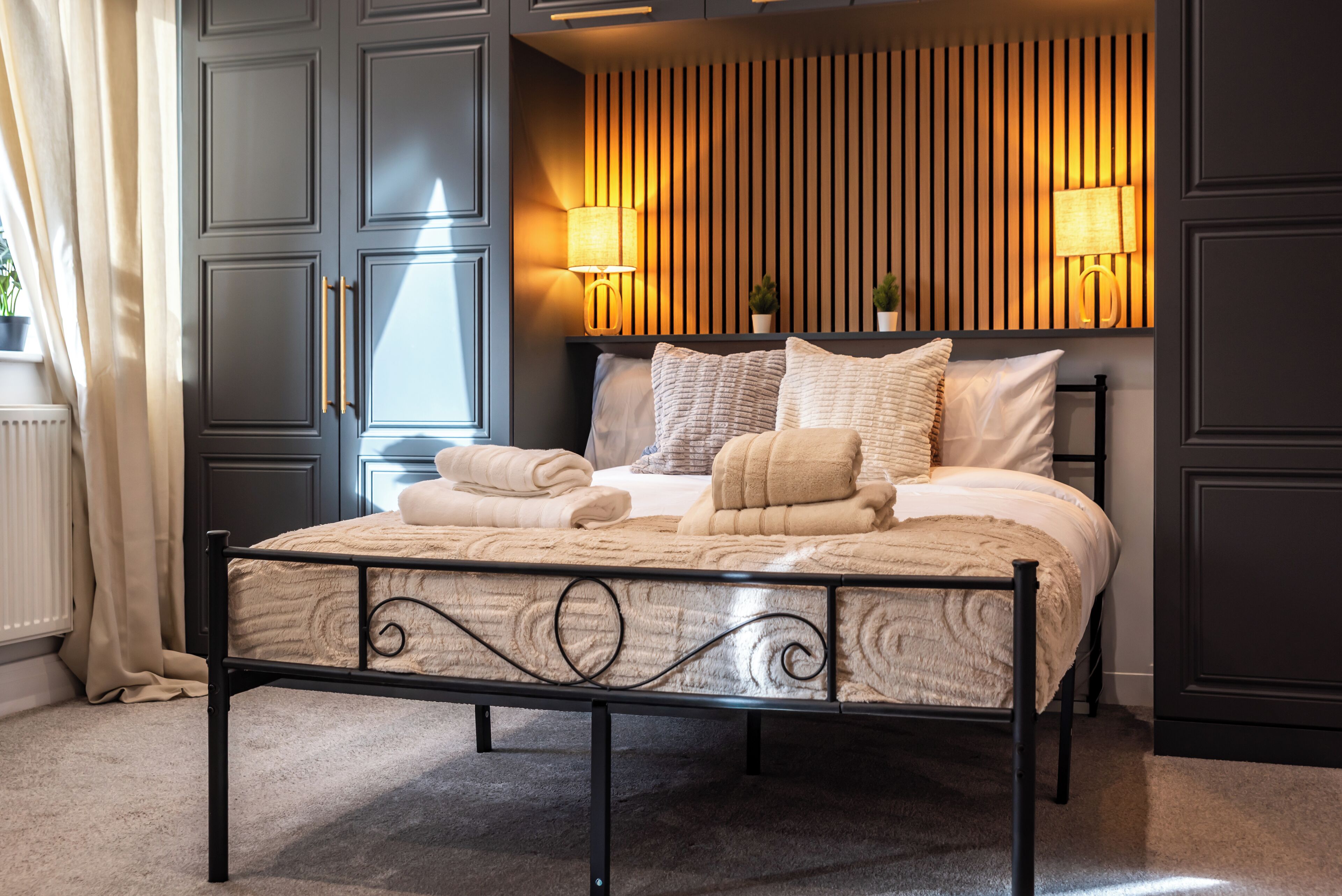 A neatly made bed with a headboard, two bedside lamps, and a window with curtains.