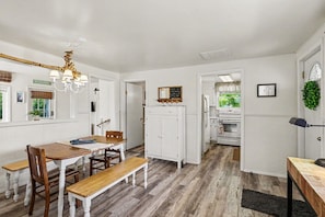 A kitchen with white cabinets, a dining table with chairs, a bench, and a hanging light fixture. - Cozy Downtown Cottage Near Beach and Pier (East Tawas)