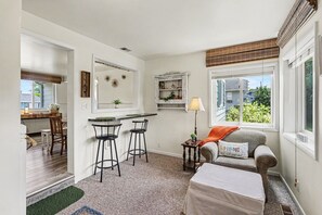 A compact living space with a kitchenette, a dining area, a sofa with a cushion, and a window with blinds. - Cozy Downtown Cottage Near Beach and Pier (East Tawas)