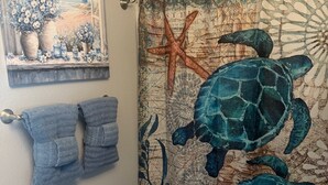 Een badkamer met een douchegordijn waarop zeeschildpadden en een zeester afgebeeld zijn, een handdoekhouder met twee handdoeken, en een klein schapje met decoratieve objecten.