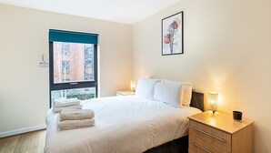 Hotellihuone, jossa on sänky, yöpöydät, ikkuna, jossa on sälekaihtimet, seinällä oleva kehystetty taulu ja näkymä ulkona oleviin rakennuksiin.
