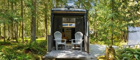 Un petit camping-car fermé, avec deux chaises à l’extérieur, entouré d’une forêt.