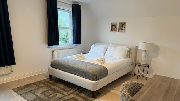 Un dormitorio ordenado con cama, mesita de noche con lámpara, una silla y una ventana con cortinas.