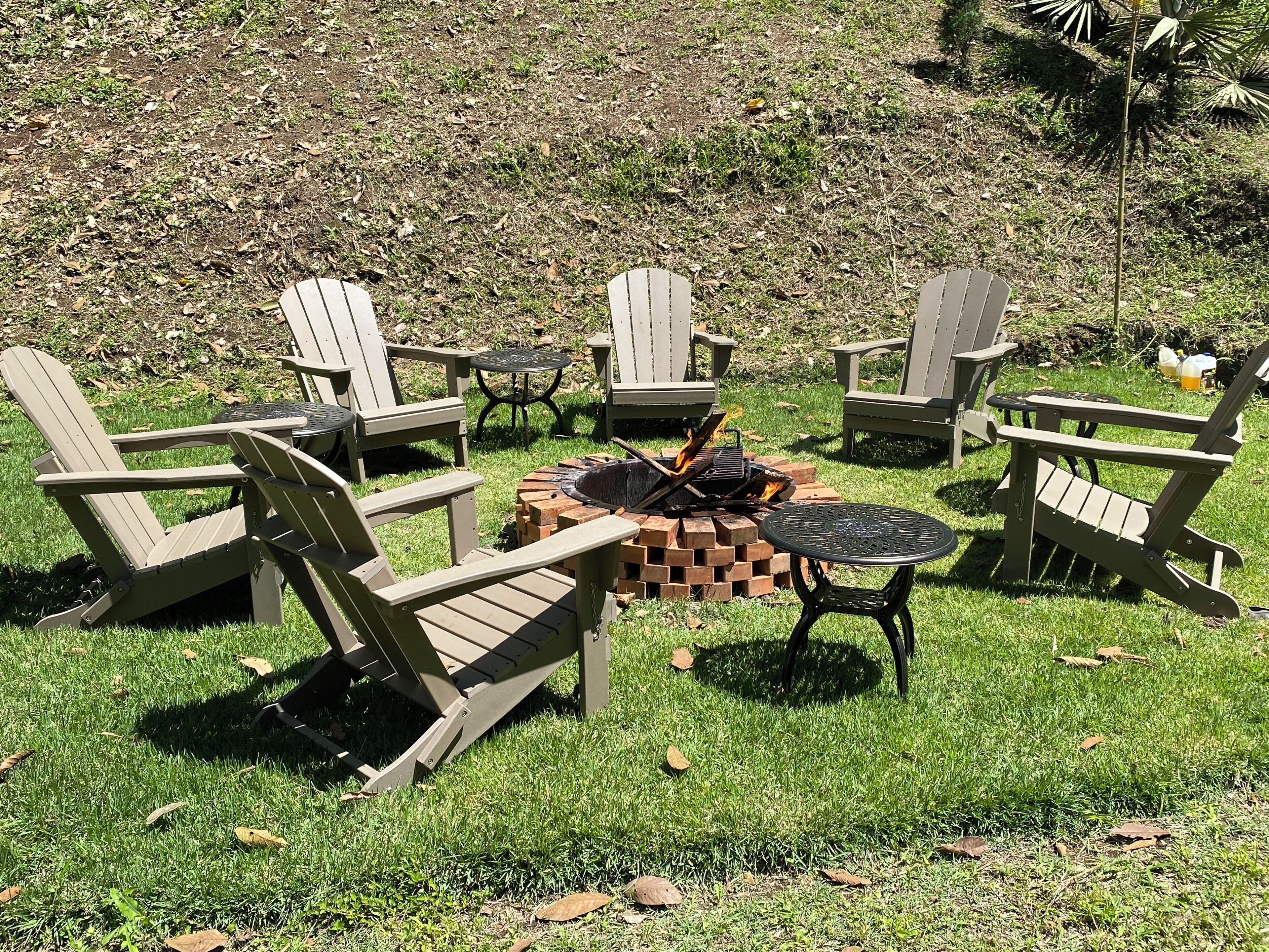 A fire pit surrounded by Adirondack chairs and a round table in a grassy outdoor area.