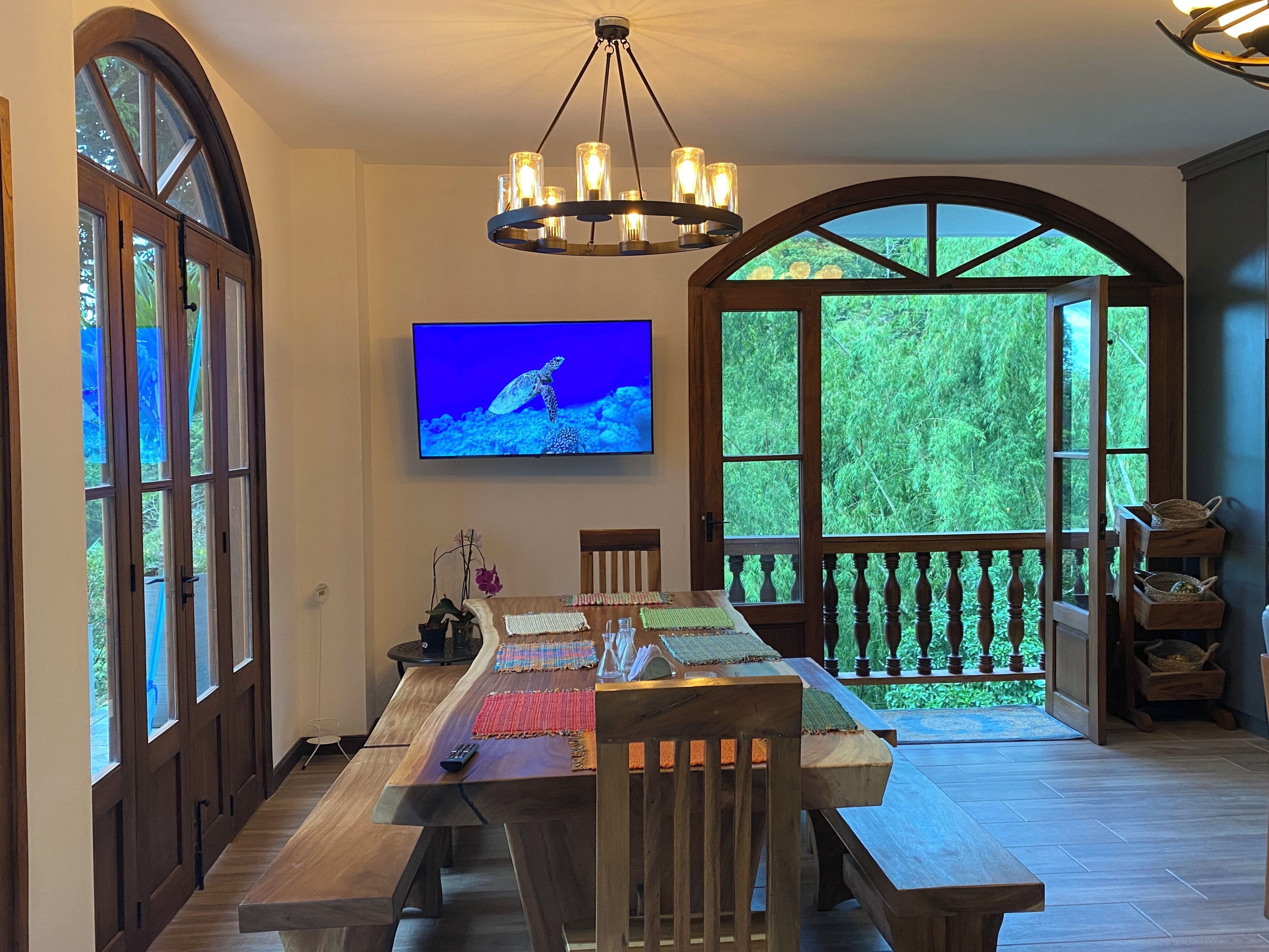 A dining area with a long wooden table, chairs, a television mounted on the wall, and large windows with a view of greenery.