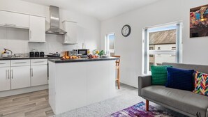 Eine moderne Küche mit weißen Schränken, einer zentralen Kochinsel und einem Essbereich mit grauem Sofa und farbenfrohen Kissen.