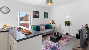 Ein modernes Wohnzimmer mit einem grauen Sofa, bunten Kissen, einem Couchtisch, einem Fernseher und einem Essbereich mit einer Theke und Frühstücksangeboten.