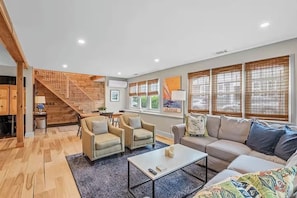 A spacious living room with a staircase, a dining area, and large windows with blinds.