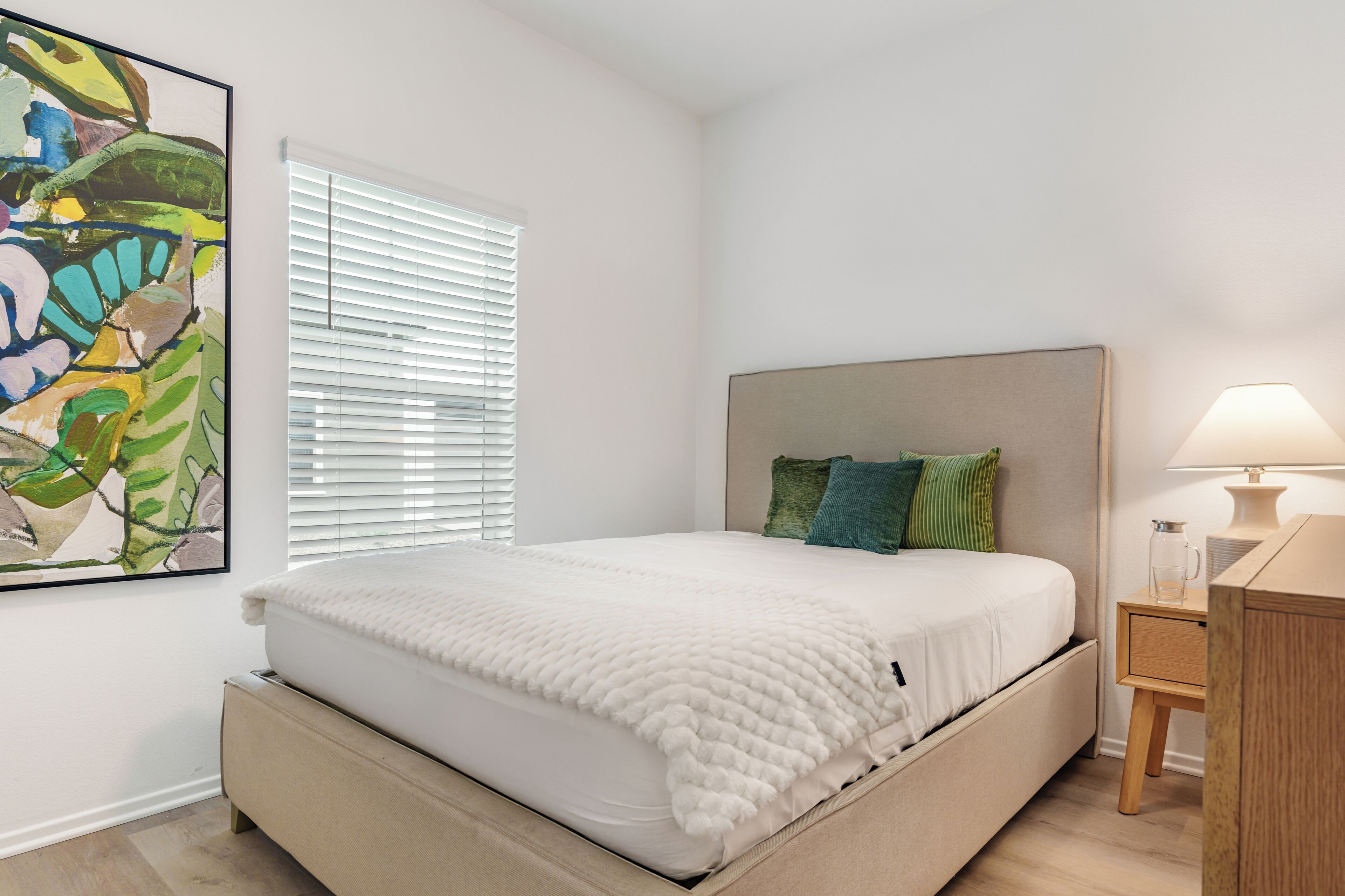 A bedroom with a bed, a nightstand, a lamp, a window with blinds, and a painting on the wall.