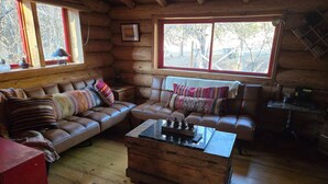 A cozy log cabin interior with a brown leather sofa, a wooden coffee table, and a large window offering a view of trees.