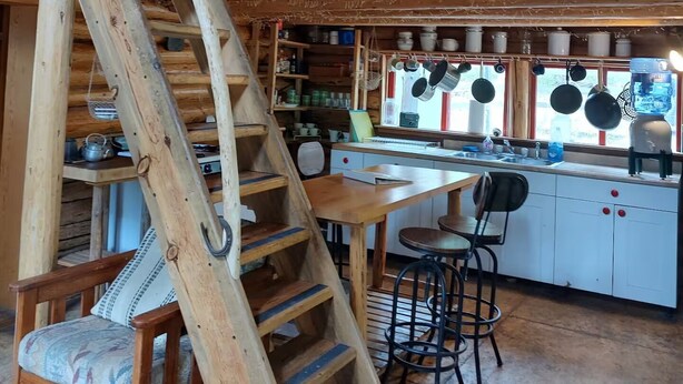 A rustic wooden cabin with a loft bedroom, a kitchen with hanging pots, and a wooden staircase.
