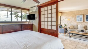 Habitación de hotel con una cama grande, un armario de madera, un televisor, una puerta corredera, un sofá, una mesa de centro y vistas a palmeras a través de la ventana.