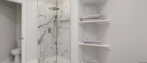 A modern bathroom with a glass-enclosed shower, white marble walls, and built-in shelves for towels.