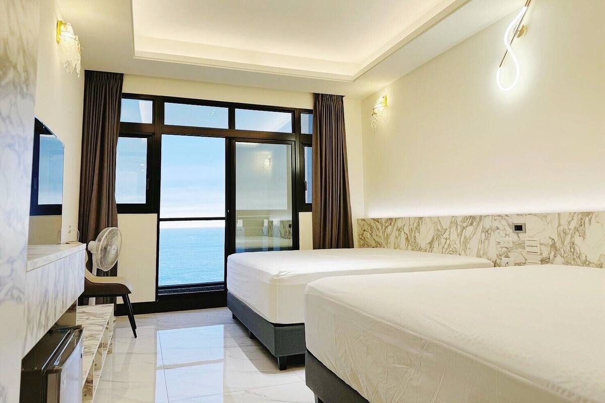 A hotel room with two beds, a sitting area with a chair, a large window with a view of the sea, and marble-patterned walls.