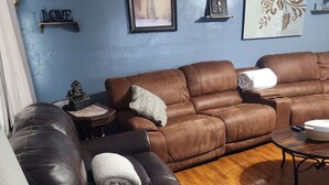 Un salon avec un canapé en cuir marron, une table basse et une table d’appoint en marbre. Des œuvres d’art encadrées ornent le mur, et une petite étagère expose des objets décoratifs.