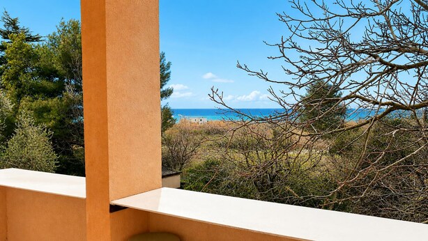 Ein Balkon mit Blick auf das Meer, ein weißer Tisch und zwei grüne Stühle.