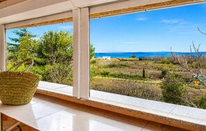 Una habitación con un ventanal grande que da a un paisaje costero, una cesta tejida en el alféizar y vistas al mar y las colinas.