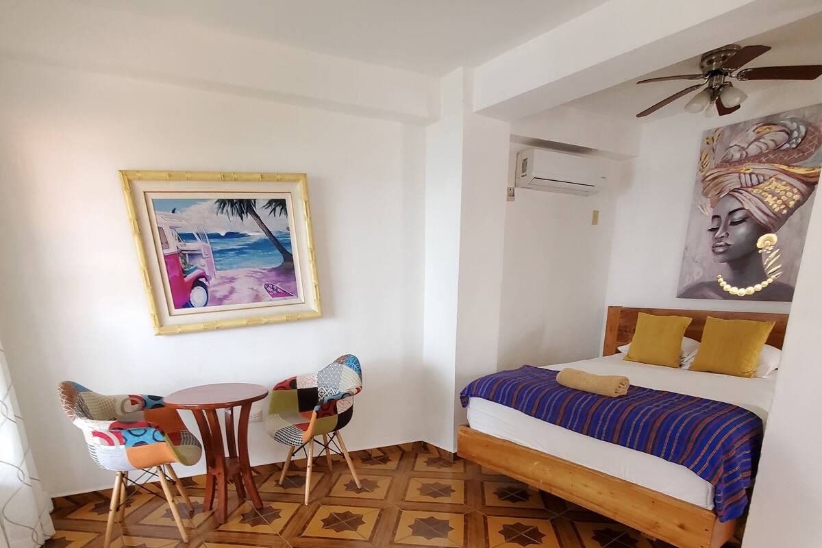 Una habitación con cama, una silla, una mesa, un ventilador de techo y un cuadro en la pared.