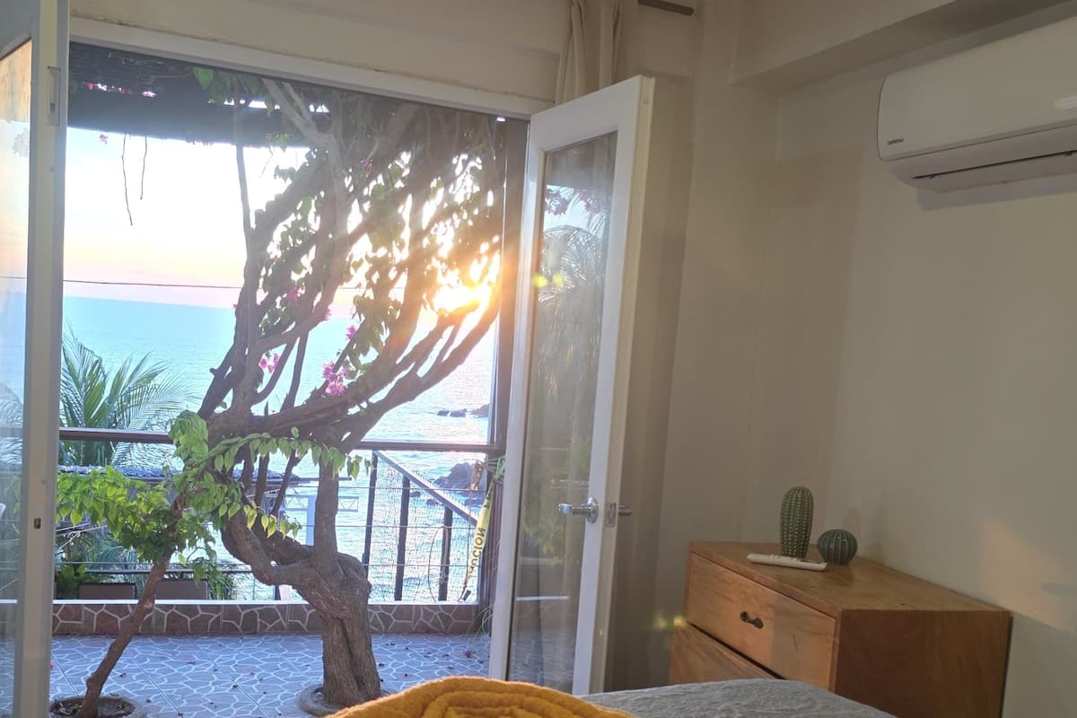 Habitación de hotel con cama, mesita de noche de madera, unidad de aire acondicionado y vista al mar a través de una puerta de vidrio.