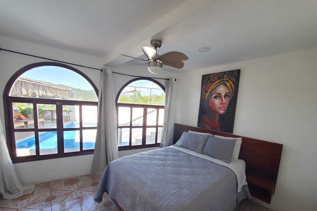 Un dormitorio con cama, ventilador de techo y amplias ventanas que dan a una piscina y zona exterior.