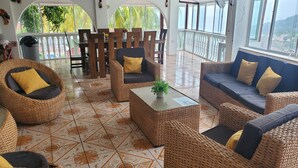 Un espacio interior amplio con mobiliario de mimbre, piso de baldosas y ventanas grandes con vistas a la vegetación.