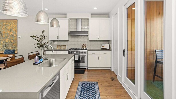 A modern kitchen with white cabinets, a central island, pendant lighting, and a view to a wooden deck. - 8th Ave South Gem Fabulous Home for Groups of 12 (Nashville)