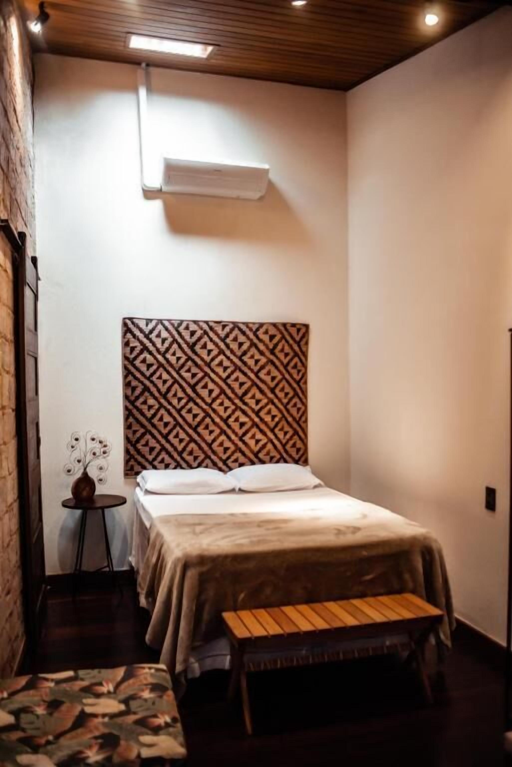 Um quarto pequeno e acolhedor com cabeceira estampada, uma cama, uma mesinha e um banco.
