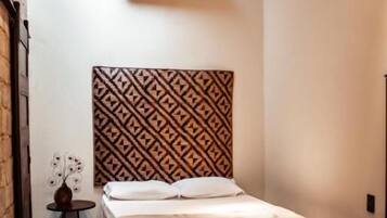 Um quarto pequeno e acolhedor com cabeceira estampada, uma cama, uma mesinha e um banco.