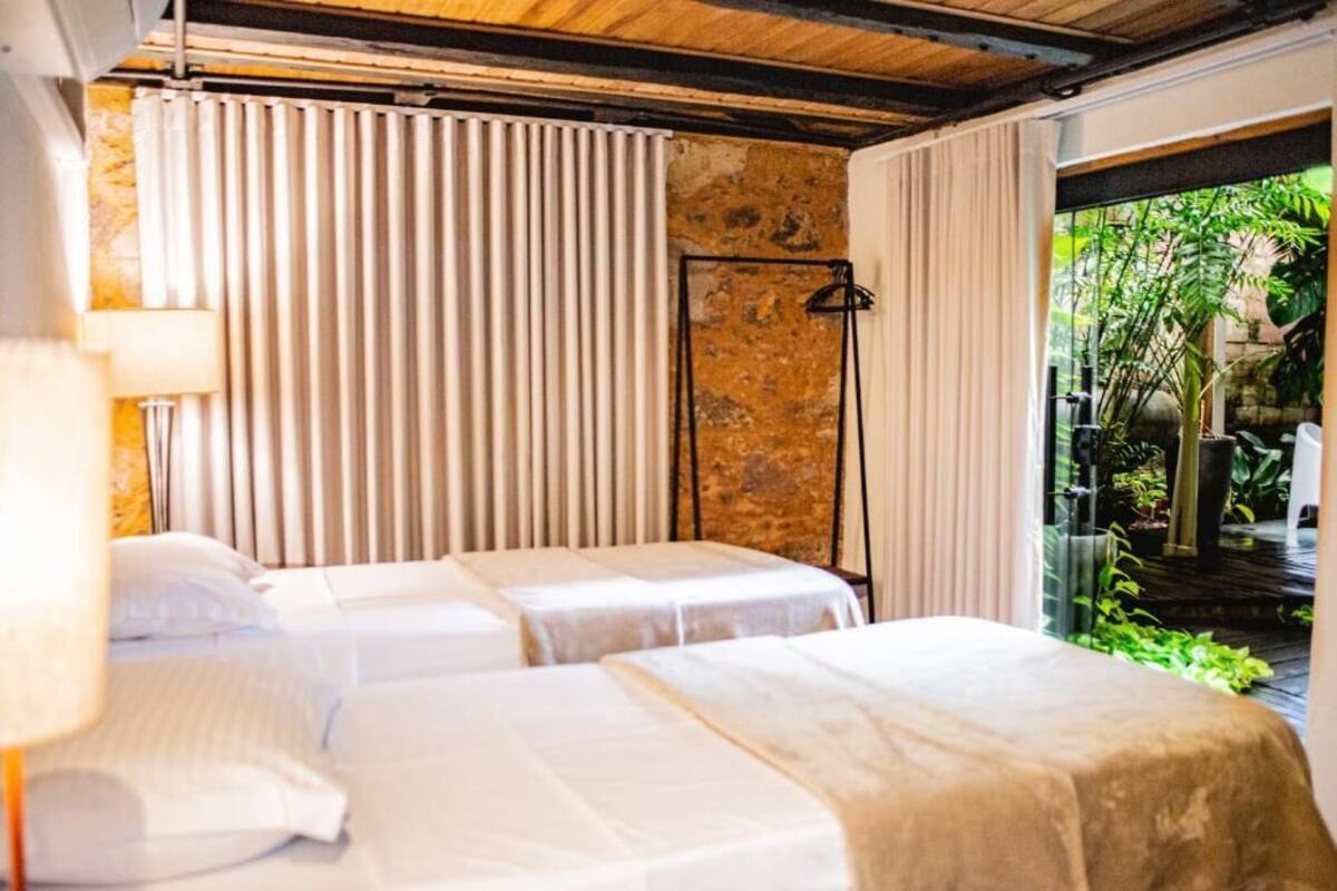 Habitación de hotel con dos camas, un ventanal con cortinas, una lámpara de pie y vistas a zonas verdes.