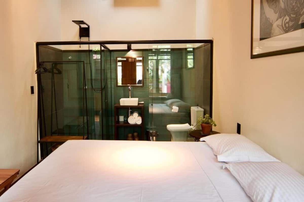 Un dormitorio moderno con cabina de ducha de vidrio, cama con ropa de cama blanca, mesita de noche con planta y una obra de arte enmarcada en la pared.
