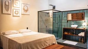 A hotel room with a bed, a bench, a glass-enclosed shower, a sink with a mirror, and framed artwork on the wall. - Cas'Amazonia Brasil (Belém)