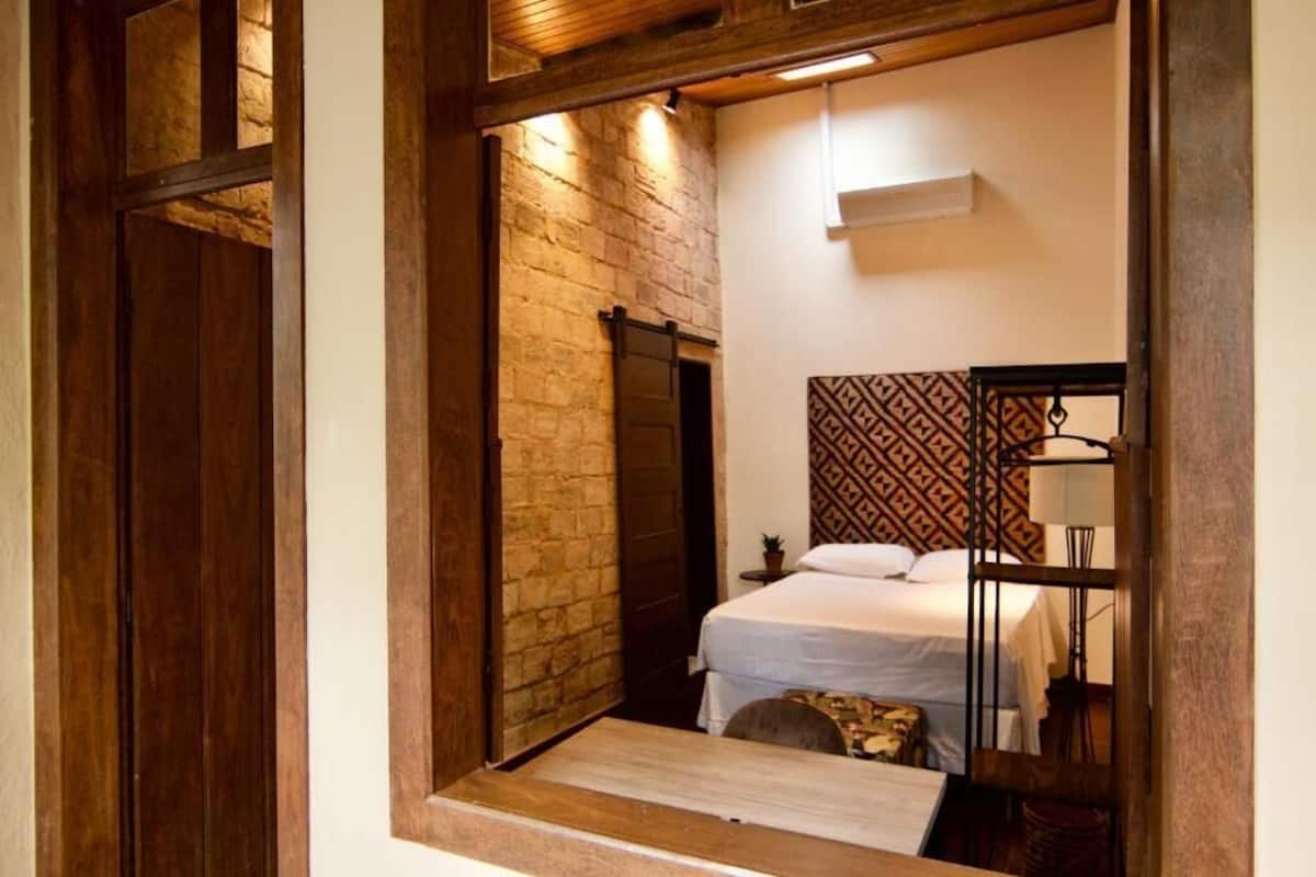 Un dormitorio con pared de ladrillo, cama con ropa de cama blanca, cabecera estampada y una mesita de noche con lámpara.